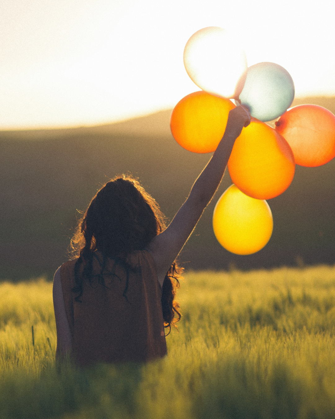 Lady in a field with her arms in the air and holding a bunch of colored balloons