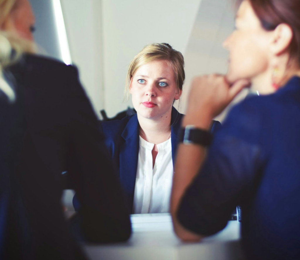 Female sitting and looking uncomfortable, opposite two other people in a workplace setting that looks like an interview or assessment