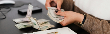 A female sitting at a desk counting money, holding dollar bills in her hand and with piles of notes on the desk