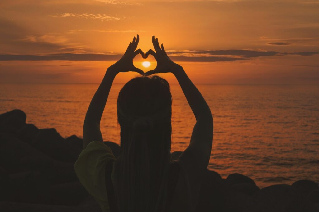 A silhouette of the back of a person standing next to the ocean with an orange sunset and they are holding their arms above their head with hands forming a heart shape and the sun is in the middle