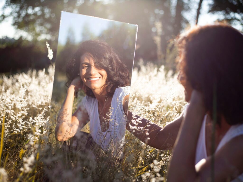 Photo of a lady sitting in a field of flowers and smiling at herself in a mirror