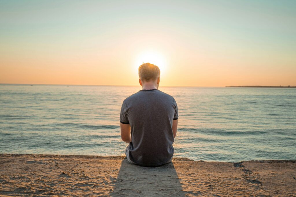 The back of a man sitting alone on a wall at the edge of the ocean at sunset
