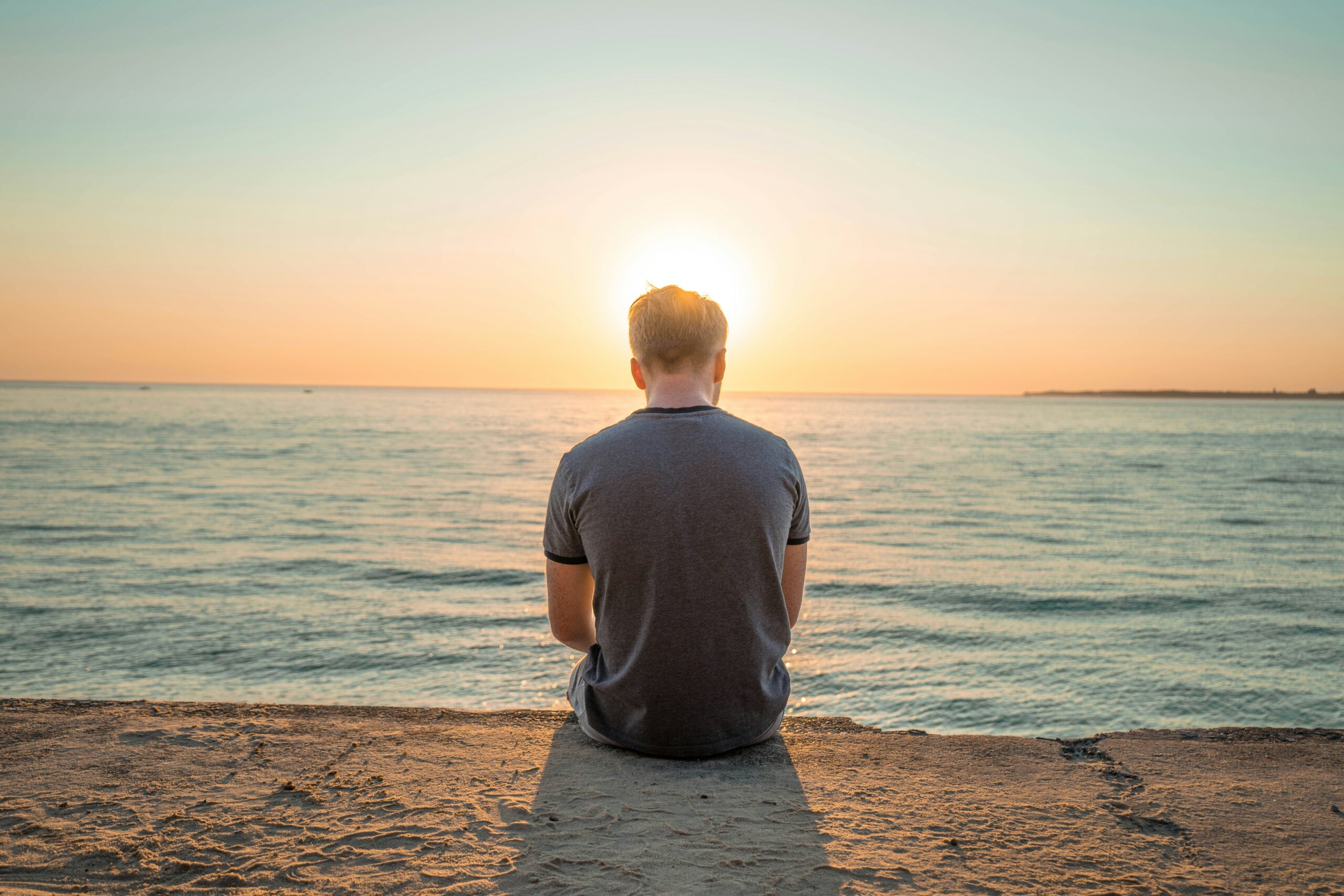 The back of a man sitting alone on a wall at the edge of the ocean at sunset