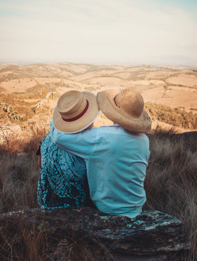 Image of the back of two people wearing hats, one with their arm around the other while sitting at the top of a hill looking over fields