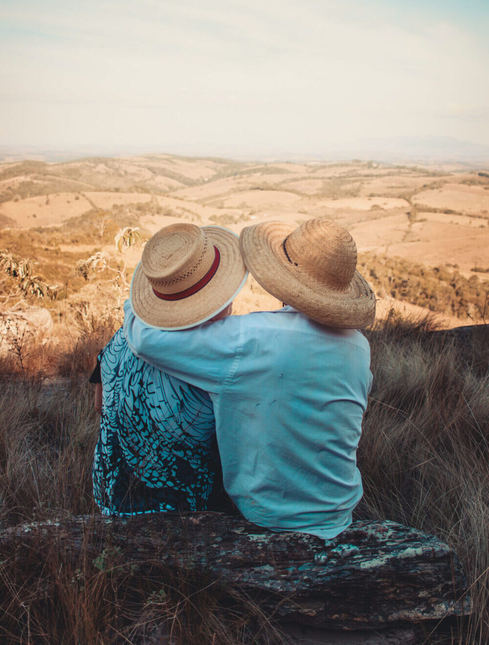 Image of the back of two people wearing hats, one with their arm around the other while sitting at the top of a hill looking over fields