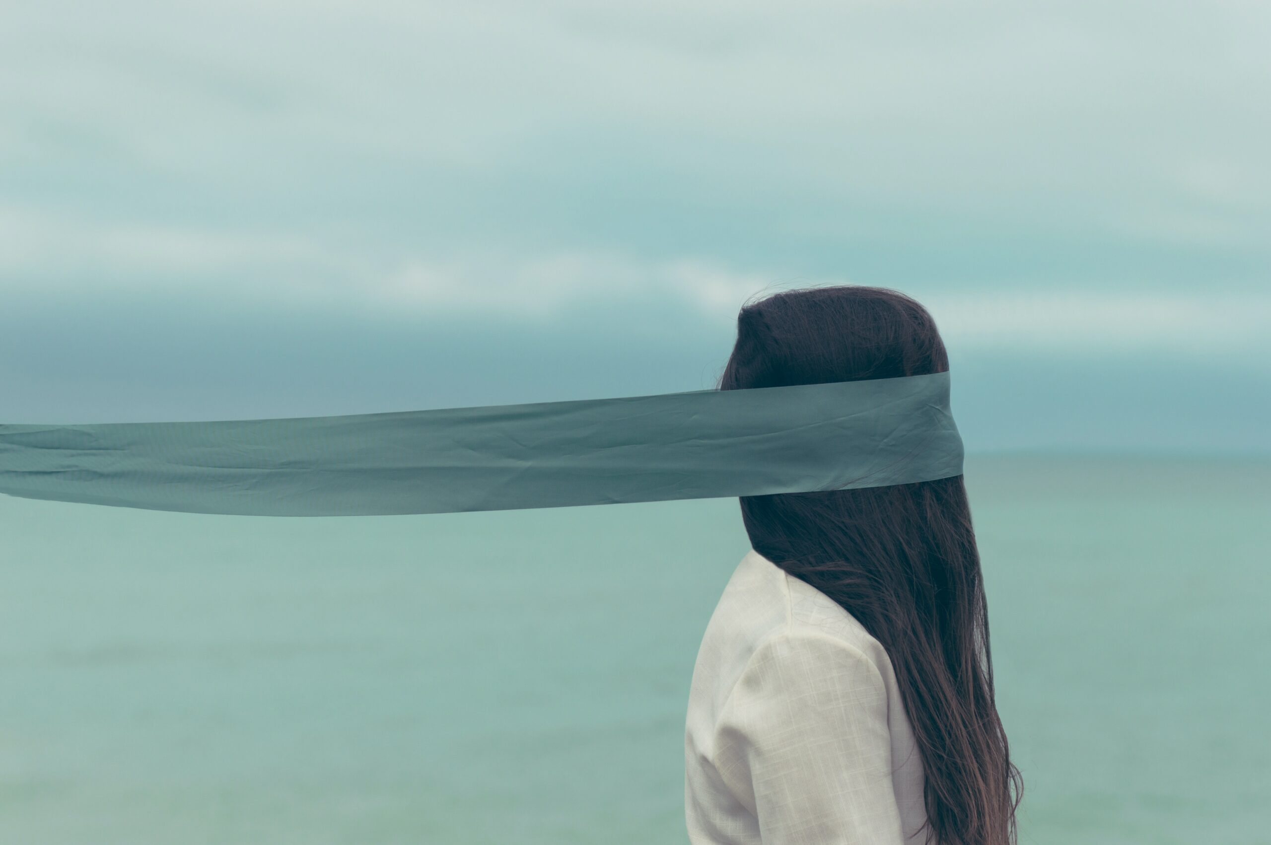 Image of a girl in front of the sea, who has a very long scarf blindfold covering her face and pulling her